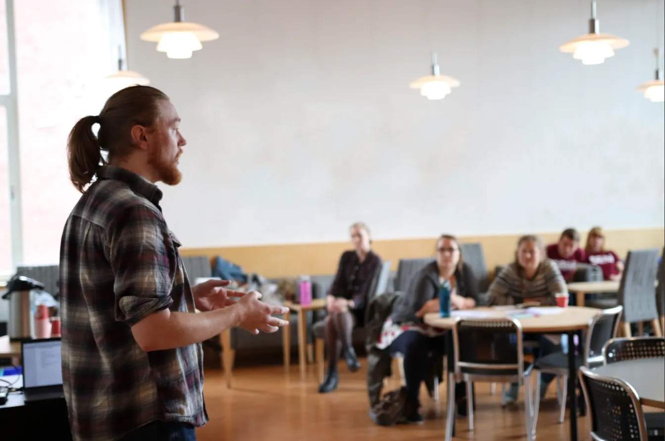 Foto av en mann med hestehale og rutete skjorte som står foran fem personer og forklarer noe med hendene ut foran seg.